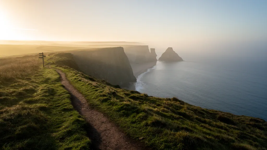 A worn grass path curving along a clifftop towards limestone headlands and a misty bay at dawn